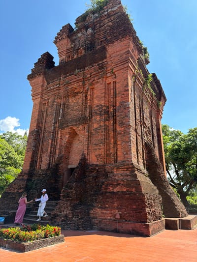 The image depicts the Cham towers of Po Nagar in Tuy Hoa, Vietnam. The ancient brick structures, weathered by time, are overgrown with vegetation, and stand against a clear blue sky. Two women are visible on the steps leading up to one of the towers. One woman, wearing a pink dress, stands on a lower step, while the other, in white clothing and a white hat, is ascending the stairs. In the foreground, a planter box filled with red flowers adds a splash of color to the scene. The ground is paved with terracotta tiles. Lush green trees surround the towers, suggesting a verdant, possibly tropical, environment.