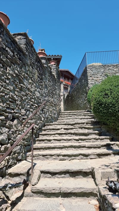A stone staircase ascends between two stone walls under a clear blue sky in Rupit i Pruit, Spain. The left wall is constructed of rough, uneven stones and features a rusted metal handrail. The right wall is similarly constructed and is topped with a black metal railing, partially obscured by a dense green bush. At the top of the stairs, a wooden building with balconies adorned with red flowers is visible. Several large terracotta pots are placed on top of the walls. The scene appears to be a historic, outdoor location.