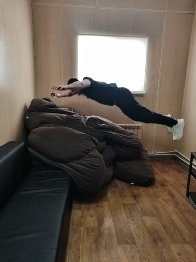 The image depicts an indoor scene in the city of Im. Gastello, Russia. An adult male is captured in mid-air, suspended horizontally across the frame. He wears a black t-shirt, black pants, and light-colored athletic shoes. His arms are extended forward, and his legs are extended backward, maintaining a rigid, elongated posture. Below the individual, a large, dark brown, heavily bundled object, resembling multiple sleeping bags or thick bedding, rests on the wooden floor against a light-colored paneled wall. To the immediate left of this brown bundle, a segment of a dark-colored sofa or bench is visible. The back wall features a rectangular window with a white frame and an opaque white covering, positioned above a white radiator and visible horizontal white piping. The floor is composed of medium brown wooden planks. A dark, multi-tiered shelf is partially visible on the far right edge of the frame.