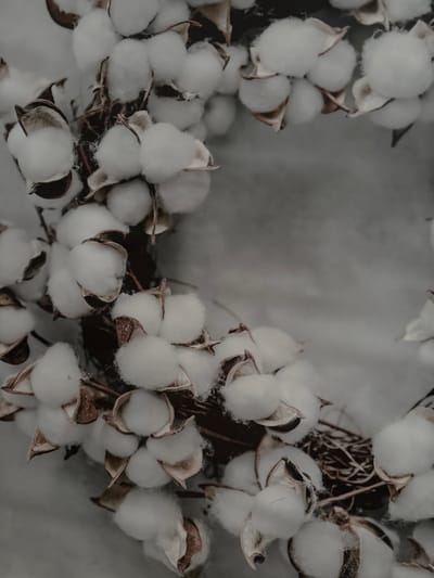 The image is a close-up, overhead shot of a wreath made from cotton bolls. The wreath is composed of numerous fluffy white cotton bolls, some still partially encased in their brown, papery husks. The bolls are clustered together, forming a circular shape, with some appearing to spill over the edges. The background is a soft, mottled gray surface, possibly fabric or a textured wall, which contrasts with the texture of the cotton. The lighting is soft and diffused, creating a gentle, muted aesthetic. The scene appears to be indoors, likely a decorative arrangement. There is no indication of the city of Imeni Morozova, Russia within the image.