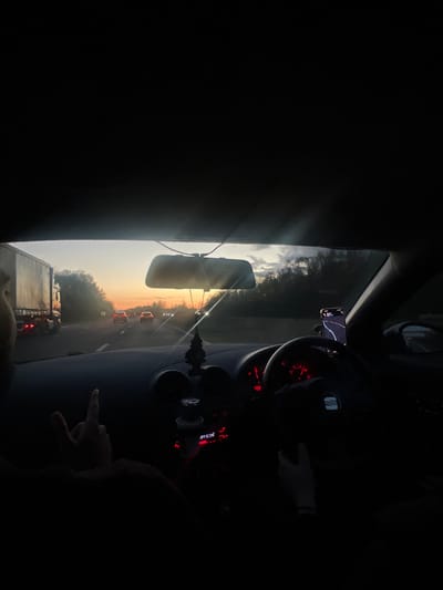 From inside a car, a view towards a highway during a bright sunset. In the rear-view mirror and through the windshield, multiple vehicles are visible on the road. The car's dashboard features a steering wheel and a phone mounted to the windshield. A hand is partially visible in the foreground, seemingly giving the middle finger. The location is indicated by a street on the dash that states “Offcial DAUNTSEY CIR."