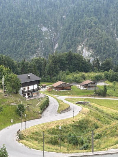 The image depicts a mountainous landscape in Sauris / Zahre, Italy, with a winding road leading past several traditional wooden houses and a larger white house with a dark roof. The houses are nestled on grassy slopes dotted with trees, and a dense forest covers the steep mountainside in the background. Several streetlights line the road. In the foreground, a group of people are seated at picnic tables near the white house.