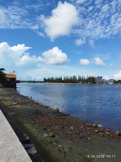 The image depicts a river scene with a sandy and debris-strewn bank in the foreground. A concrete walkway borders the left side of the frame. Across the blue water, a tree-lined shore is visible with several buildings, including what appears to be a hotel or multi-story apartment complex. The sky is a clear blue with scattered white clouds. A date and time stamp, "18.06.2026 09:11", is present in the lower right corner of the image. This scene is located in Kampot, Cambodia.