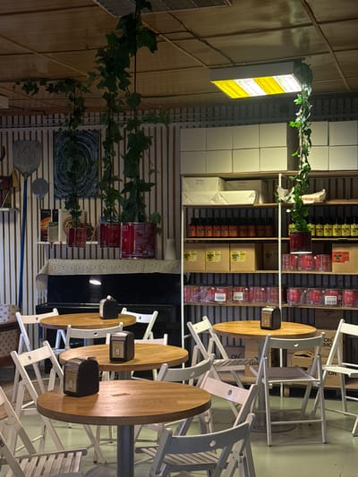 The image depicts an indoor scene, likely a restaurant or cafe, with a rustic ambiance. The setting features wooden paneling on the walls and shelves filled with various items, including what appear to be bottles of olive oil and canned goods. A prominent element is a bright, rectangular light fixture with alternating yellow and white panels, suspended from the ceiling and partially adorned with green ivy. Several wooden tables and white folding chairs are arranged throughout the space, with some tables stacked and chairs folded, suggesting a space that can be reconfigured. One table is set with a small black dispenser. On one wall, there's a piece of abstract art with swirling blue and green patterns. The overall impression is that of a casual, perhaps somewhat dimly lit establishment, possibly in the evening given the lighting. There is no visible indication of people or weather. Some text is visible on the labels of the products on the shelves, such as "OLIVE OIL" and "BOSSO".