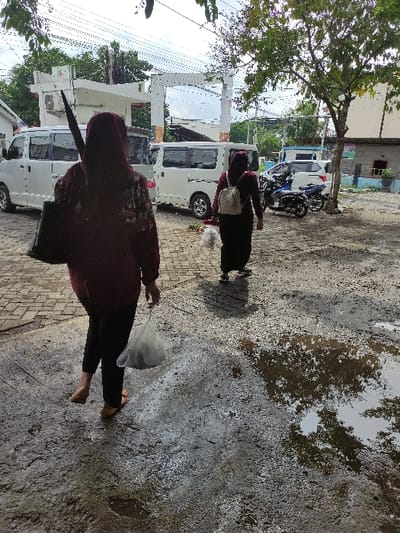 This media file captures an outdoor street scene in Kecamatan Asembagus, Indonesia, during the daytime. The image is taken from a slightly elevated perspective, looking down a street.
Two individuals are the primary subjects, seen walking away from the camera. The person in the foreground, on the left, wears a dark, long-sleeved top and dark pants, appearing to be barefoot. They are carrying a white plastic bag in their right hand. Further down the street, a second person, also wearing dark clothing and a light head covering (possibly a hijab), walks in the same direction.
The ground surface is a mixture of paving stones and uneven, wet earth with numerous puddles. These puddles reflect the bright sky and overhead trees, suggesting recent rainfall.
In the background, several white vans or minibusses are parked along the street. Behind them, parts of buildings are visible, including a white, columned structure. Parked motorbikes can also be seen further down the street, nestled between vehicles and buildings. A tree with green foliage is prominent on the left side of the frame, providing shade. The overall setting is an urban or semi-urban environment with traffic and pedestrian activity. No visible text is discernible in the image.
