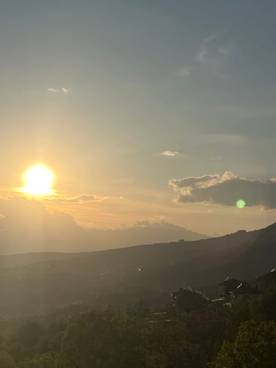 The image depicts a sunset view from Kribul, Bulgaria. A bright sun is positioned low on the horizon, partially obscured by clouds. To the right, a mountain range is visible, with some houses and lush green vegetation at its base. The sky above the sunset displays varying shades of blue, transitioning to golden hues closer to the sun. A scattering of clouds is present in the sky.