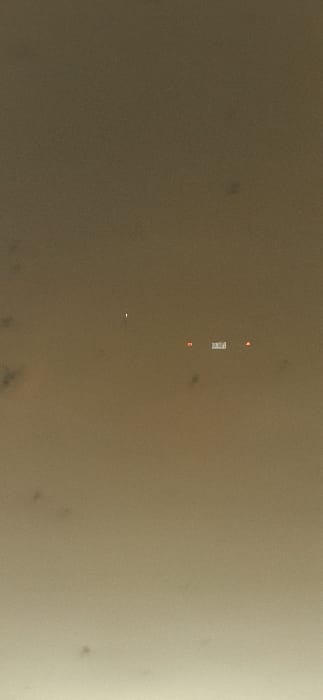 A blurry, tan-colored aerial view shows a group of objects. A rectangular, light-colored object is centered, flanked above and below by small, red rectangles. A small, bright, white spot is located below the main grouping. The image has a grainy texture with darker smudges scattered across the background, especially in the bottom left corner. The right side of the image is slightly lighter than the left.