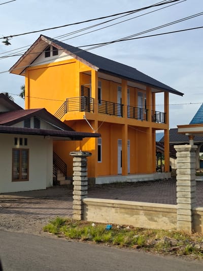 The image shows a bright orange, two-story building with a dark roof, located in Ierhobglumpang, Indonesia. The building has several windows and balconies with black metal railings. To the left of the orange building, there is a smaller, cream-colored building with a brown roof. A paved driveway separates the buildings, and in the foreground, there is a road and some grassy areas with scattered debris. Power lines are visible across the sky, and the overall scene suggests a residential or institutional area. There are no people visible in the image.