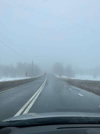 The image displays a two-lane asphalt road in Isakovo, Russia, viewed from inside a vehicle's dashboard perspective. Heavy fog and an overcast sky obscure the distant horizon. The road surface is wet, reflecting ambient light, with a double solid white line separating the left lane and dashed white lines marking the right lane. Snow and dirt accumulation lines both road shoulders, with puddles evident along the right side. Multiple utility poles with streetlights and overhead power lines are visible along the left side of the road, receding into the fog. Bare trees are discernible through the dense fog on both sides of the roadway. In the distance, an oncoming red heavy vehicle, likely a truck or bus, is partially visible, followed by a smaller white vehicle, both significantly obscured by the fog. No individuals or explicit actions are present in the scene.