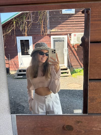 A mirror reflects a young woman taking a selfie with a smartphone. She wears a leopard print bucket hat, sunglasses, a light pink, cropped, holey sweater, and white high-waisted pants. The reflection includes the facade of a two-story wooden building with white-framed windows and an air conditioning unit mounted on the wall. The environment suggests a residential area in Aya, Russia, based on typical building styles and street surfaces found in that region.