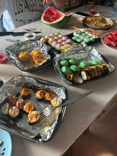 The image depicts a table laden with various food items, suggesting a meal or gathering. In the foreground, several black foil containers hold portions of sushi rolls. One container features sushi with a crispy, fried exterior, some topped with a creamy green sauce and drizzled with dark sauce. Another container contains sushi rolls with a golden-brown, breaded coating, some topped with a lighter creamy sauce and what appears to be roe. A third container, also lined with foil, holds several sushi rolls partially consumed, with one roll being lifted by a fork. There's a small dish of soy sauce, a knife, and a pink condiment visible.

Further back on the table, a slice of watermelon is prominent, as is a partially eaten pie in a round dish. Another plate holds cut pieces of watermelon. The table surface has a subtle patterned texture. In the background, part of a person's torso is visible, wearing a grey t-shirt with "LOS ANGELES CITY OF ANGELS" printed on it. The setting appears to be indoors, possibly a dining area. The overall impression is of a casual meal with an assortment of food. The location of Патруши, Russia is not depicted or implied by any elements within the image.