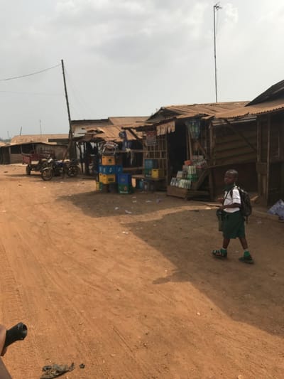 The scene depicts a rural street in Ogbese, Nigeria, under an overcast sky. A young male child, dressed in a white short-sleeved shirt, green shorts, green socks, and sandals, walks right-to-left across a reddish-brown dirt road. He wears a dark backpack and carries a green bag in his left hand. To the child's left, a row of market stalls constructed from wood planks with corrugated metal roofs lines the street. In front of these stalls, stacks of plastic crates, some bearing "Heineken" branding in blue, yellow, and green, are present. A display stand holds numerous bottled beverages. Further left, two motorcycles are parked next to a small, red two-wheeled trailer. A tall, slender pole with wires extends from the left side of the frame, and a similar antenna-like structure is visible further back on the right. Scattered debris and litter are visible on the ground. A dark object, possibly part of a bicycle or a hand, is partially visible in the bottom left foreground, suggesting the photographer's perspective.