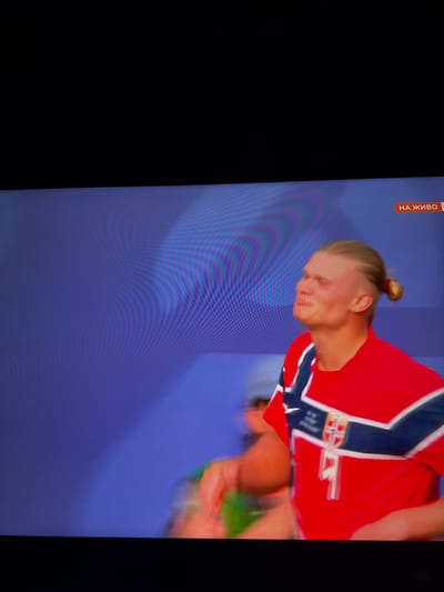 In a dimly lit room, a man with blonde hair pulled back in a bun is shown from the chest up, facing right and looking upwards with his eyes closed and mouth slightly open. He is wearing a red jersey with blue and white stripes across the chest, and a crest on the left side of his chest. A small orange rectangle with Cyrillic text "НА ЖИВО" is visible in the top right corner of the frame. The background is a blue, textured surface with faint horizontal lines. The image appears to be a screen capture from a broadcast.