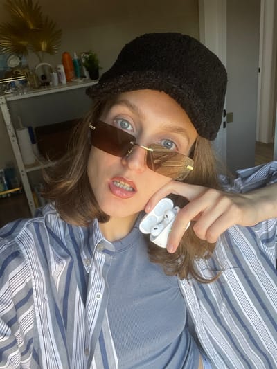 The image is a selfie-style photograph of a young woman. She is wearing a black knit hat, brown-tinted sunglasses with a gold frame, and a striped collared shirt over a blue-gray t-shirt. She is holding a white charging case with two wireless earbuds in her right hand near her face. The background reveals a room with shelves displaying various objects and a decorative gold palm frond. The location is unspecified, but the user provided Sepych, Russia as a possible location.