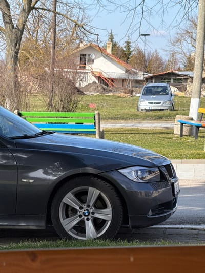 The scene depicts a daytime exterior view in Drandar, Bulgaria. In the immediate foreground, the front quarter of a dark grey BMW vehicle, appearing to be a 3-series (E90/E92 generation), is visible. Its front wheel, bearing a BMW logo on the hubcap, headlight, and a portion of the kidney grille are discernible. The vehicle is situated on a dark paved surface next to a narrow strip of green grass. A wooden element or railing occupies the bottom of the frame.
In the midground, a grassy area features sparse deciduous trees with bare branches, indicative of late winter or early spring. Two park benches with concrete bases are present: one on the left with green and blue slats, and another on the right with multi-colored slats, partially obscured by a light-colored utility pole. A paved walkway or street separates the foreground vehicle from these benches.
The background is dominated by a two-story white residential building with a reddish-brown tiled roof and a chimney. An external orange-red staircase ascends to an upper-level entrance, and a satellite dish is mounted on the wall. Piles of disturbed earth or construction debris are visible near the building's foundation. To the right of this residence, a silver minivan, consistent with an Opel model (e.g., Zafira), is parked on uneven ground within a grassy expanse. Its Bulgarian license plate, "B 3404 HB," is clearly visible. Behind the minivan, a smaller, rustic outbuilding or shed can be seen. The sky overhead is clear and blue. No individuals are present in the image.