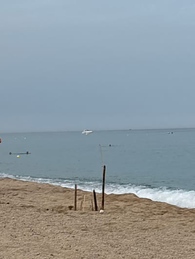 The image depicts a beach scene in Vilassar de Mar, Spain, on a hazy day. The foreground is dominated by sandy beach with some weathered wooden stakes partially buried in the sand. The sea occupies the middle ground, with gentle waves breaking near the shore. In the water, a white speedboat is visible in the distance to the right, and several indistinct figures appear to be swimming or floating, one of them near a yellow buoy on the left. The horizon line is blurred by the overcast sky, which is a uniform light gray.