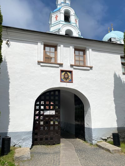 The image shows the entrance gate to a monastery in Sortavala, Russia. The gate is made of dark metal and has an arched opening. Above the gate is a white building with two windows and a mosaic of Jesus' face. In the background, a white bell tower with a blue roof and a blue dome topped with a golden cross can be seen. The sky is blue with some clouds. The area around the gate is paved with cobblestones and there are some trees and bushes.