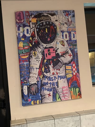 The image presents a colorful, collage-style artwork featuring an astronaut as the central figure. Bold, graffiti-like words and phrases like "LUXGMOON," "SUPER STAR," "FIRST MAN ON THE MOON," "FAST LIFE," and "MONEY PLANET" are scattered around the astronaut. The composition incorporates a variety of elements, including numbers, stars, cartoonish images, and corporate logos, creating a visually busy and layered aesthetic. The artwork appears to be a blend of pop art, street art, and commercial imagery.