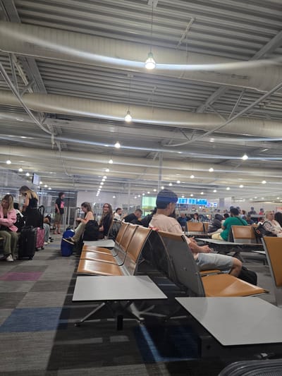 The image shows an interior view of an airport terminal in Spata-Loutsa Municipal Unit, Greece. The scene is a waiting area with rows of seats, some occupied by passengers. Luggage is visible near the seats and in pathways. The ceiling features exposed ductwork and lighting fixtures. In the foreground, a man in a light pink t-shirt and denim shorts is seated, facing away from the camera. In the background, other passengers are seen sitting or standing, with a departure/arrival information screen visible further back. The overall environment suggests a busy airport concourse.