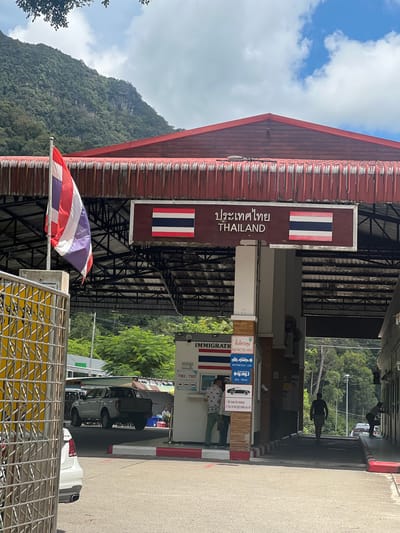 The image depicts a border crossing between Thailand and Malaysia, specifically at Wang Kelian. The foreground features a Thai flag, and a sign with "ประเทศไทย THAILAND" is visible on a building with a red, corrugated roof. Further into the scene, an immigration checkpoint is present, indicated by signs with "IMMIGRATIO" and Thai text, along with pictograms for motorcycles, cars, buses, and trucks. There are also instructions for travelers: "1. Roll down the windows 2. Turn on the light inside your car." Several people are seen at the checkpoint, some appearing to be officials and others travelers. Cars and trucks are lined up, suggesting ongoing border activity. The background shows lush green mountains and trees under a bright, cloudy sky, indicating it's daytime with fair weather. The overall impression is a functional and active border crossing point.