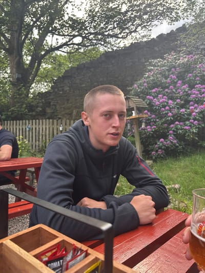 The image shows a young man sitting at a red picnic table in a garden setting in Hayfield, United Kingdom. He is wearing a black fleece jacket with a red stripe on the sleeve. His arms are crossed in front of him, and he is looking directly at the camera. Behind the man is a stone wall and a patch of bushes with purple flowers. A wooden fence is also visible, and another person is sitting at a separate table in the background. A wooden box with condiment packets is on the table in front of the man. A glass of beer is partially visible in the lower right corner of the image. A wooden birdhouse with a metal roof can be seen mounted on the wall. The man is sitting on a red wooden bench and there is grass on the ground around the picnic tables. The sky is partially visible in the background, but it is mostly obscured by the tree. The image is likely taken on a sunny day.