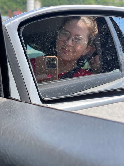 A person's reflection is visible in a car's side-view mirror. The person is wearing glasses and has their hair pulled back. They are holding a smartphone, which is taking the picture. The mirror is slightly dirty with water spots. The car appears to be silver. In the background, out of focus, are some green trees and a blue sky. The scene is likely in Bakovići, Montenegro, as indicated by the prompt. The foreground shows the interior of the car, including a portion of the door panel.