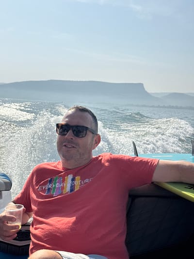 A male individual is depicted in a moving recreational boat on the Sea of Galilee, near Migdal, Israel. The Caucasian male, approximately 30-40 years old, has short dark hair and stubble, wears dark sunglasses and a red short-sleeved t-shirt with a "EVERYDAY COUTURE" graphic featuring vertical colored stripes. He is seated, holding a clear plastic cup containing an amber liquid in his left hand. His right arm rests on a padded backrest. Behind him, boat spray and wake are visible on the water surface. A yellow-green surfboard or wakeboard with a fin is positioned to his right on the boat's interior. In the background, a body of water extends to the horizon, with hazy, undulating mountains or hills visible under a clear, bright sky.