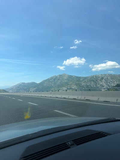 The image captures a daytime view from inside a vehicle traveling on a highway. The road is asphalt with white lane markings. A concrete barrier with metal railings separates the highway from the adjacent landscape. In the background, a range of rugged, green mountains under a bright blue sky with scattered white clouds dominates the horizon. The foreground shows the dashboard of the vehicle, including a windshield wiper and part of the car's interior. The scene is likely located near Bolesestra, Montenegro, given the characteristic mountainous terrain and highway infrastructure often found in the region. There are no people or specific actions visible in the image, it is a static landscape shot.