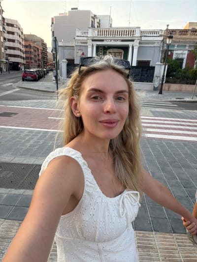 A young woman with blonde hair, wearing a white eyelet dress and star earrings, is taking a selfie on a street in Benimàmet-beniferri, Spain. She is standing on a pedestrian crossing with red and white stripes. Behind her, a street is lined with buildings and parked cars. A building with the name "VILLA MARIA" is visible in the background. The sky is clear, suggesting it is daytime. The woman is holding hands with another person, whose hand is visible on the right side of the frame.