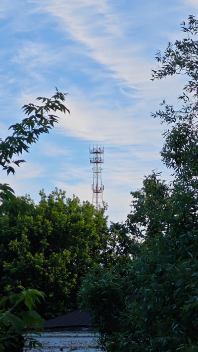 The image shows a cell tower against a blue sky with wispy clouds. The tower is tall and metallic, with various antennas and equipment attached. It is framed by lush green trees on either side, with some leaves in the foreground appearing slightly out of focus. In the lower portion of the image, a building with a dark roof and white brickwork is partially visible, partially obscured by trees. The scene appears to be an outdoor, possibly rural or suburban, setting.