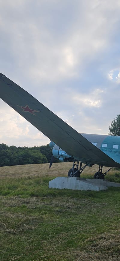 In the town of Vyšný Komárnik, Slovakia, a vintage aircraft, a Soviet-era Ilyushin Il-14 transport plane, is displayed outdoors. The plane is painted in a camouflage scheme of dark green and blue, with a red star insignia visible on the port wing. It is mounted on concrete stands in a grassy field, with a mowed field and a line of trees in the background. The sky above is cloudy, with patches of sunlight breaking through. The aircraft appears to be in good condition, likely preserved as a monument. There are no people visible in the scene.