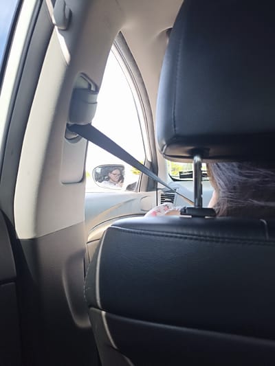 The image shows a view from inside a car, looking towards the rear passenger seat and the side window. A person with dark hair wearing glasses is reflected in the side mirror. The person is wearing a light-colored shirt with a floral pattern. The interior of the car is visible, including the black leather headrest of the front passenger seat and the beige pillar with a seatbelt. The scene suggests the car is moving, indicated by the slight blur in the reflection and the context of being inside a vehicle. The image was taken in the city of Dungloe, Ireland.