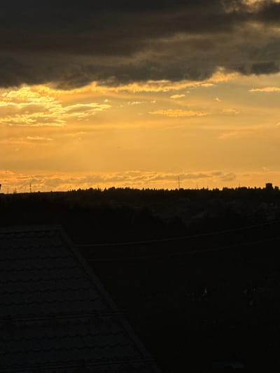 The image captures a twilight scene in Pychanki, Russia. The primary subjects are the sky and a dark, silhouetted landscape. The sky is dominated by hues of yellow and orange, suggesting either a sunset or sunrise, with streaks of lighter clouds catching the light. To the left, darker, more opaque clouds create a stark contrast. The horizon line is marked by a dense line of trees, appearing as a dark, jagged silhouette against the lighter sky. Several thin, vertical structures, possibly utility poles or antennas, are visible in the distance along the treeline. To the far right of the image, a dark, textured surface, likely the roof of a building, slopes downwards. The overall mood is serene and atmospheric, typical of dusk or dawn. There are no people or discernible objects in the foreground. The time of day is clearly evening or early morning. The weather appears to be clear apart from the clouds, and there are no visible text elements.
