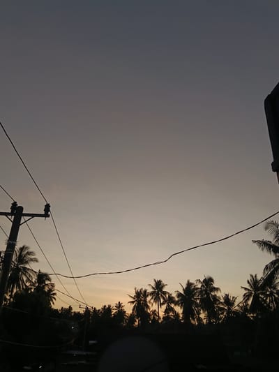 The image captures an outdoor scene in Lebbae, Indonesia, at either dawn or dusk, characterized by a gradient sky and silhouetted elements. The upper sky is a dark, desaturated blue-grey, transitioning downward to a lighter grey-purple, and then to a faint orange-yellow hue at the horizon, indicating the presence of a light source below the visible frame. Minimal cloud cover is apparent. A dark utility pole, equipped with insulators and crossarms, is positioned on the left side of the frame, from which multiple dark electrical wires extend horizontally and diagonally across the sky. In the upper right corner, a dark, rectangular architectural structure is partially visible. The mid-ground is dominated by a dense, horizontal line of numerous silhouetted palm trees, their distinctive fronds outlined against the lighter horizon. The immediate foreground, beneath the tree line, consists of indistinct dark shapes, including a large, dark, dome-like object in the lower center. No individuals or specific actions are depicted within the scene.