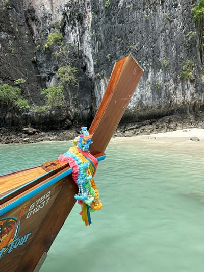 A longtail boat is approaching a rocky island covered in green foliage. The boat's bow is decorated with a lei of colorful flowers. The water is a clear turquoise. The background shows a sandy beach and more of the rocky island. The boat has registration numbers "6752" and "01237" on its side, along with the words "Tour."