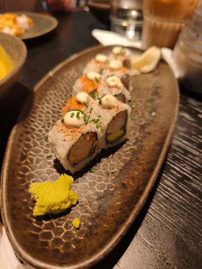 A close-up, eye-level shot captures a plate of sushi rolls and wasabi on a dark, textured surface, likely a wooden table in Bnei Dror, Israel. The sushi, presented on a dark, oval plate with a honeycomb pattern, consists of several maki rolls. Each roll is topped with a dollop of white sauce, sprinkled with red seasoning and chopped green chives. A small mound of vibrant green wasabi sits on the left side of the plate. In the blurred background, other plates of food and drinks suggest a dining setting. The lighting is dim, creating a warm and inviting atmosphere.
