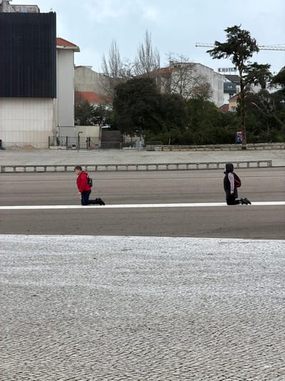 The image depicts an outdoor scene at the Sanctuary of Fátima, Portugal, under an overcast sky. Two individuals are kneeling on a dark asphalt esplanade, positioned on or near a distinct white line that runs horizontally across the midground.
The individual on the left, an adult female, wears a red jacket, dark trousers, and dark shoes, with a black backpack on her shoulders. She is kneeling on her right knee, facing slightly to the right of the frame. The individual on the right, smaller in stature, wears a dark hooded top, a dark vest, dark trousers, and dark shoes, with an orange backpack. This person is kneeling on both knees, facing the same direction as the first individual.
The immediate foreground consists of a textured, light-colored cobblestone pavement. In the background, various urban and natural elements are visible. To the left, a large modern building with a dark, vertically ribbed facade stands adjacent to a white building featuring a red-tiled roof. Further right, a multi-story grey building displays a red "HOTEL" sign. Scattered trees, some appearing sparse or with broken branches, are situated between the esplanade and the background structures, separated by low metal barriers. A construction crane is partially visible above the "HOTEL" building on the horizon.