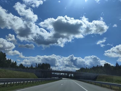 The image depicts a highway passing under a bridge in Skujnieki, Latvia. The sky is a vibrant blue, filled with dramatic white and gray cumulus clouds. Sunlight streams from the upper right corner. A paved road, with white lane markings, stretches into the distance. On either side of the road are metal guardrails and grassy embankments. A concrete bridge spans the highway, with trees and a dense forest visible beyond it. The bridge has a railing along its top. The scene is a daytime outdoor shot, likely taken from a moving vehicle, capturing a common view of Latvian infrastructure and natural landscape. There are no people or animals visible in the image.