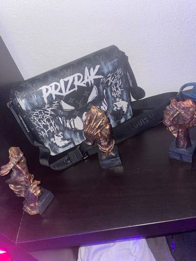 An indoor scene displays various items arranged on a dark, flat surface, likely a table or shelf. The primary subject is a black Louis Vuitton shoulder bag, customized with white graffiti-style art. The word "PRIZRAK" is prominently stenciled in white across the front of the bag, above a stylized white face or mask featuring two eye cutouts and a jagged mouth. Additional white text, including repeated "HAHAHA" patterns and ghost-like motifs, covers the bag's surface. The bag's black strap, partially revealing "LOUIS VUITT" branding, extends from beneath it to the right.
Positioned in front of the bag are three small, bronze-colored figurines, each mounted on a dark rectangular base. From left to right: the first figurine depicts a head with hands covering the eyes; the central figurine portrays a pair of hands clasped together; and the third, on the right, is a head or bust.
The items rest on a dark, possibly polished wood surface that shows minor scratches. The background consists of a plain, light-colored wall. A purple light source is visible in the lower left of the frame, casting a purple hue on the immediate foreground and beneath the surface. No individuals are present. The image contains no visual information to determine a specific location within 's-Hertogenbosch, Netherlands.