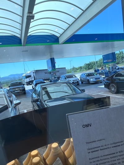 This media file shows a scene at a gas station, likely in Kalugerovo, Bulgaria, given the location context. The perspective suggests the photo was taken from inside a vehicle.
The primary subjects are several cars lined up at the pumps. One car, a silver sedan with license plate "27-SP-XV", is prominently visible. Behind it, a black SUV with license plate "CB 3122 P8" is also in line. Further back, a white truck with a dragon graphic on its side is visible. There are no people clearly identifiable in the image.
The setting is a covered gas station forecourt. The blue and green branding of "MAXX Motion" and "OMV" is visible on signs, indicating the brand of the station. A sign with Bulgarian text, reading "Уважаеми клиенти, Уведомяваме ви, че от съображения за се приемат банкноти с номинал от Б Възможни са отклонения от 0,01 по касова бележка, превалутирана за конкретна стока или усп подбите на За", is also present. This text appears to be a customer announcement regarding payment with banknotes and potential discrepancies.
The scene appears to be set during the daytime, with bright sunlight creating reflections on the vehicles. The sky is clear and blue. The overall atmosphere is that of a typical, busy gas station.