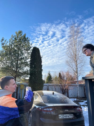 The image depicts an outdoor scene in Заозерное Снт, Russia, under a clear blue sky with scattered cirrocumulus clouds. The ground is partially covered with snow and bare earth.
Two individuals are present. In the left foreground, a male, young adult with short dark hair, wearing an orange, blue, and black windbreaker over a light-colored hoodie, stands with his left profile visible. His right arm is raised, bent at the elbow, with his hand clenched into a fist, gesturing upwards and slightly to the right. A blue glove or sleeve is on his right hand. He exhibits a slight smile and looks towards the right.
In the right mid-ground, partially obscured by a dark, vertical structure, another male, young adult with dark hair and glasses, is visible from the shoulders up. He wears a light-colored, possibly plaid, long-sleeved shirt. He leans forward slightly, looking downwards and to the left, towards the center of the frame.
A dark-colored Kia Stinger sedan is parked in the mid-ground, facing away from the viewer. Its rear license plate, "P354PH 198 RUS," is clearly legible. The rear window and trunk exhibit a light dusting of what appears to be snow or frost.
The background features various trees: a large evergreen conifer on the far left, a tall, slender evergreen conifer centrally located behind the car, and several bare deciduous trees, including a birch tree with visible white bark, extending above the right individual. A wooden slatted fence runs across the mid-background. A yellow gas meter box is affixed to a wooden post behind the left individual. Sunlight is prominent, casting visible shadows.