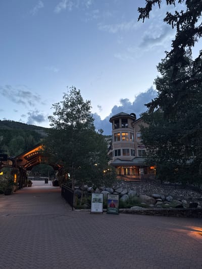 This is an outdoor, eye-level, daytime, vertical, natural light photograph of a town square in Beaver Creek, United States of America. In the foreground, a brick-paved walkway leads through an arched wooden structure adorned with lights and signage. Beyond the arch, the walkway continues toward a multi-story building with a distinctive round tower. The building has several floors with numerous windows, some of which are lit. The surrounding environment includes lush green trees, rocky landscaping, and a mountain in the background. The sky is a gradient of blue and gray with scattered clouds, indicating dusk or dawn. Signs are visible along the walkway, including one for "CRANIALOGIE" and another advertising a "CREEKSIDE PARK CONCERT SERIES." Another sign visible on the archway reads "THANKS." No people are clearly visible in the photograph.