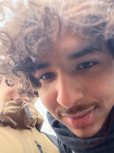 This close-up image, purportedly taken in Meistratzheim, France, captures two individuals outdoors. The foreground, occupying the right side of the frame, shows an individual with medium-dark skin, dark curly hair, and a mustache, looking directly at the camera with a closed-mouth smile. This person wears a dark, ribbed scarf. Partially visible behind and to the left of the foreground subject is a second individual. Only the upper portion of this second person is discernible, including curly hair, a section of their face showing the chin and mouth, and the collar and upper chest of a light brown jacket featuring silver grommets and dark zippers. The background is a bright, out-of-focus expanse with faint outlines of bare tree branches.