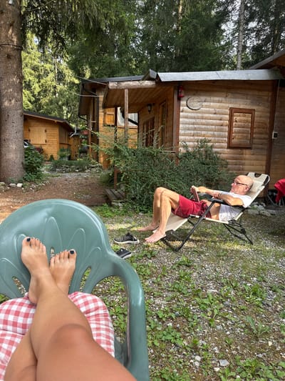 An outdoor scene in a wooded area with several rustic wooden cabins. In the foreground, a person's legs are visible, crossed at the knees, resting on a red and white checkered cushion on a green plastic chair. The person's feet, with dark nail polish, are propped up. To the right, a man is relaxing in a white and tan reclining chair, wearing glasses, a white t-shirt, and red shorts. He is looking at a tablet in his hands. A pair of black sneakers lies on the gravel ground near him. The environment suggests a campsite or a secluded vacation spot, likely in or near Borno, Italy, given the natural setting and wooden structures.