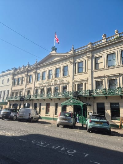 The image depicts the Green Dragon Hotel situated on Broad Street in Hereford, United Kingdom, under a clear, bright blue sky.
The prominent structure is a multi-story hotel building with a light-colored facade, likely beige or cream, featuring classical architectural details. The name "GREEN DRAGON HOTEL" is visible on the upper facade. A Union Flag, red, white, and blue, flies from a flagpole atop the central pediment, supported by a dark, possibly green, dragon statue. The building exhibits numerous rectangular windows, some with dark frames, and decorative green ironwork balconies on the first floor. A distinctive green ornamental canopy covers the main entrance at ground level.
On the asphalt street in front of the hotel, an inverted and partial "LOADING" road marking is visible. Several vehicles are parked along the curb. From left to right: a dark grey hatchback, a white panel van, a silver hatchback, and a light blue/mint green compact SUV. The silver hatchback and the light blue/mint green compact SUV are parked directly in front of the hotel's main entrance.
One individual, partially obscured by the light blue/mint green vehicle and the hotel canopy, is visible near the hotel entrance. They appear to be wearing light-colored clothing, possibly blue or green, and are positioned near the doorway. Sunlight casts shadows from unseen objects onto the street in the foreground. Overhead wires are discernible against the clear sky.