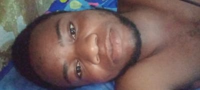 The image displays a close-up, horizontally oriented view of an adult male in an indoor setting in Mairi, Nigeria. The subject has dark skin, short dark hair, and a dark, short beard. His dark eyes are open and directed towards the camera, maintaining a neutral expression. He appears to be shirtless, with bare skin visible on his right shoulder and upper chest. He is positioned lying down on a blue fabric, which exhibits a pattern of yellow, pink, and light blue circular or amorphous shapes in the upper left background. To the immediate left of his head, a lighter, textured surface, possibly a wall or another fabric, is visible with minor discoloration.
