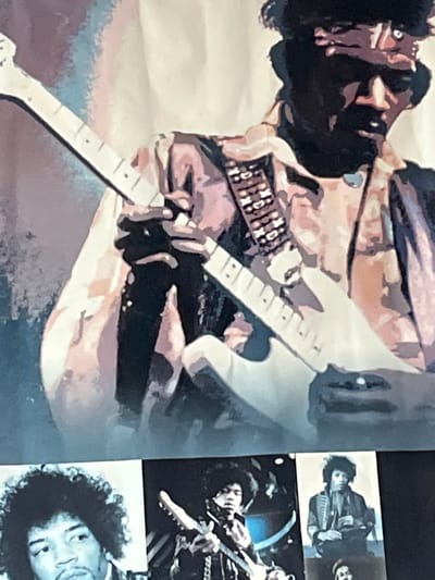This multi-part display, located in Ancenis, France, predominantly features musician Jimi Hendrix. The upper two-thirds of the scene show a large, stylized graphic of Hendrix playing a white electric guitar. He is depicted from the chest up, facing right and slightly downward, wearing a dark bandana, a collared shirt, and a guitar strap adorned with distinct, light-colored rectangular embellishments. His left hand is positioned on the guitar's fretboard, while his right hand is partially visible near the guitar's body, appearing to interact with the strings. His face is largely obscured by shadow, revealing a prominent mustache and goatee. The graphic utilizes a block-color aesthetic with shades of brown, pink, purple, black, and white against a lighter, subtly colored background with hints of blue. The lower third of the display presents three distinct black and white photographic prints arranged horizontally. The leftmost photograph is a headshot of Hendrix looking left. The central photograph depicts Hendrix playing an electric guitar on a stage. The rightmost photograph shows Hendrix in patterned attire, potentially seated, with an indistinct object held in his hands; a partial fourth image is visible below it.