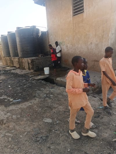 This image depicts a dusty street scene in Gobirawa, Nigeria, under bright daylight. In the mid-ground, a row of at least four large, black cylindrical water storage tanks stands on a raised concrete platform. Adjacent to these tanks is a light-colored building with a single rectangular window fitted with horizontal bars.

Three adult males are positioned near the water tanks. One male, wearing a red short-sleeved shirt and dark shorts, stands next to a white bucket, appearing to be engaged with a water tap connected to the tanks, suggesting water collection. To his right, another male in a white short-sleeved shirt and a dark beanie leans against the building wall. A third male, partially obscured, stands behind the individual in the white shirt, also near the wall.

In the foreground and mid-ground, three children are walking from right to left across the frame. The most prominent child, positioned centrally, wears a light orange long-sleeved tunic and matching trousers, with light-colored slip-on shoes. To his immediate left and slightly behind him, a second child is partially visible, wearing a blue shirt. Further to the right, a third child, also dressed in a light orange tunic and trousers, walks with dark sandals on his feet.

The ground is unpaved, consisting of dry earth and scattered debris, including plastic and paper. A partially covered concrete drainage ditch or gutter runs across the mid-ground, separating the walking children from the adults and water tanks. The scene suggests daily activities related to water access and pedestrian movement in the community.