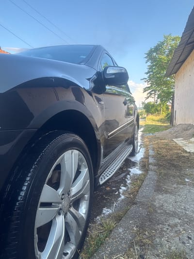 The image shows the side of a black car with a silver wheel and a running board. The car is parked on a dirt and gravel surface next to a concrete curb with some grass. There is soapy water on the ground and on the running board, suggesting the car is being washed. In the background, there is a building with a corrugated metal roof and a tree. The sky is blue with some clouds, and there are some power lines visible. The overall scene appears to be an outdoor residential area. The location is confirmed to be in the city of Katselovo, Bulgaria.