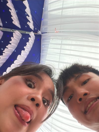 The media file presents a playful, low-angle portrait of two individuals, likely young adults, looking down towards the camera. The scene is set in Tay Son Commune, Vietnam.
The person positioned higher in the frame, closer to the top edge of the image, displays a cheerful expression with a visible smile and teeth. They have short, dark hair. Directly below, the face of a second individual is prominently featured, characterized by wide, expressive eyes and their tongue playfully sticking out. Their dark hair frames their face, adding to the dynamic composition. Both subjects convey a lighthearted and humorous mood.
The background is distinctly divided. The upper section consists of soft, sheer white drapery or curtains, suggesting an airy or illuminated space. The lower section reveals a deep blue fabric or canopy, adorned with numerous small, twinkling blue lights that create a starry effect. White, ruffled fabric decorations are horizontally arrayed against the blue backdrop, contributing to a festive atmosphere. A metallic support structure is also visible within the decorated background.
The setting appears to be an indoor or covered outdoor venue, elaborately decorated for a celebratory event such as a party or gathering, possibly in the evening given the presence of decorative lighting. The photograph captures a candid and intimate moment between the individuals and the camera. No visible text is present in the image.